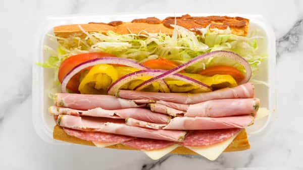 Fresh deli sandwich loaded with layers of ham, salami, and provolone cheese, topped with shredded lettuce, tomato slices, banana peppers, and red onion on a toasted sub roll, served in a takeout container.