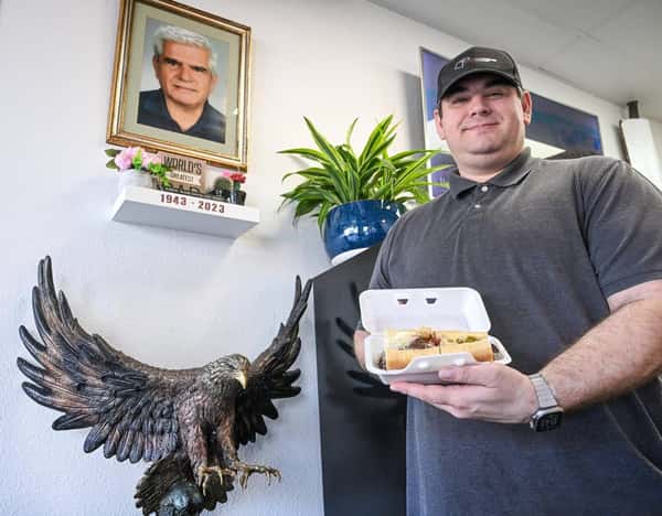 Man holding a sandwich in a takeout box inside Eagle Sandwich Co., standing beside a wall with a bronze eagle sculpture and a memorial shelf featuring a framed portrait, flowers, and a sign reading "World's Greatest" with dates 1943–2023.