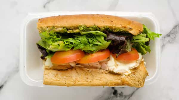 Fresh chicken pesto sandwich with leafy greens, sliced tomatoes, creamy mozzarella, and a layer of vibrant pesto sauce on toasted artisan bread, served in a takeout container.