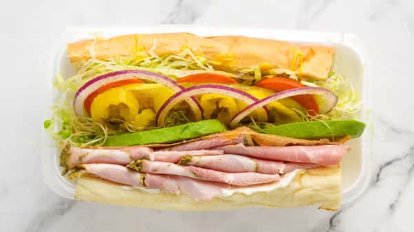 Fresh deli sandwich with layers of sliced ham, creamy avocado, shredded lettuce, tomato, banana peppers, and red onion on a soft hoagie roll, served in a takeout container.