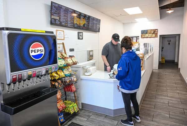 Inside Eagle Sandwich Co., a customer in a blue jacket places an order at the counter while a staff member assists. A Pepsi soda fountain, shelves stocked with assorted chips, and a large menu board are visible in the casual deli setting.