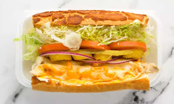 Buffalo chicken sub sandwich with melted cheese, shredded lettuce, tomato slices, banana peppers, and red onion on a toasted hoagie roll, served in a takeout container.