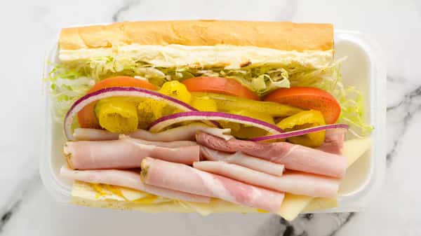 Fresh deli sub sandwich with layers of ham, Swiss cheese, shredded lettuce, tomato slices, banana peppers, and red onion on a soft hoagie roll, served in a takeout container.