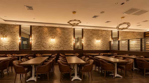 Restaurant dining room with leather chairs and wooden tables against a stone wall, illuminated by modern chandeliers.