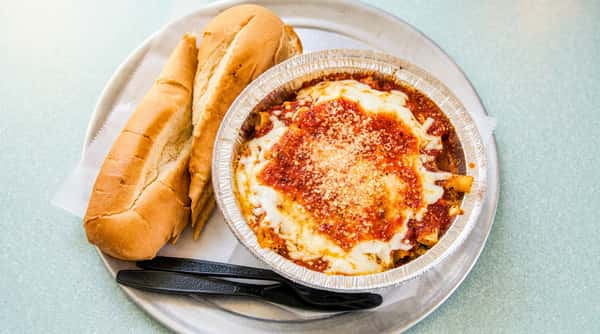 Baked ziti in a foil pan with melted mozzarella, marinara, and Parmesan, served with two baguette halves.