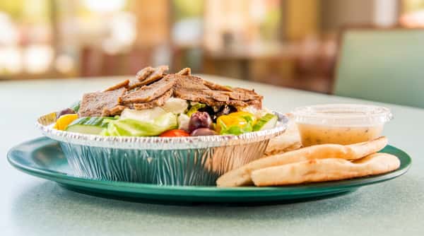 Gyro platter in foil container with sliced meat, lettuce, cucumbers, tomatoes, kalmata olives, feta, pita bread, and dressing.