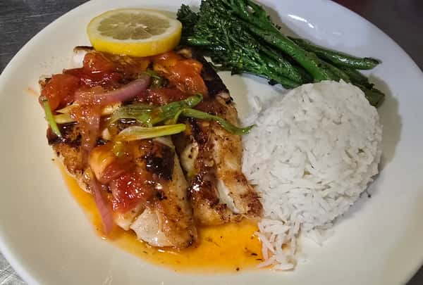 Grilled fish fillet topped with sautéed vegetables and served with white rice, broccolini, and a lemon slice.