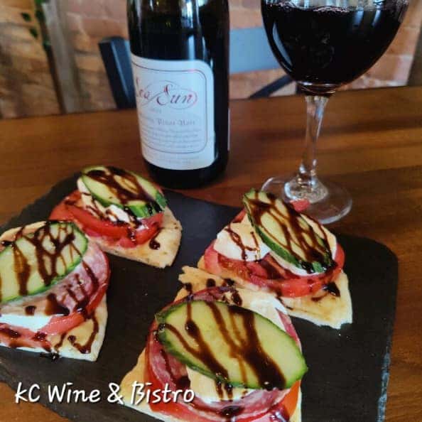 Gourmet flatbread slices topped with cucumber, tomato, mozzarella, salami, and balsamic glaze, served with a glass and bottle of red wine.