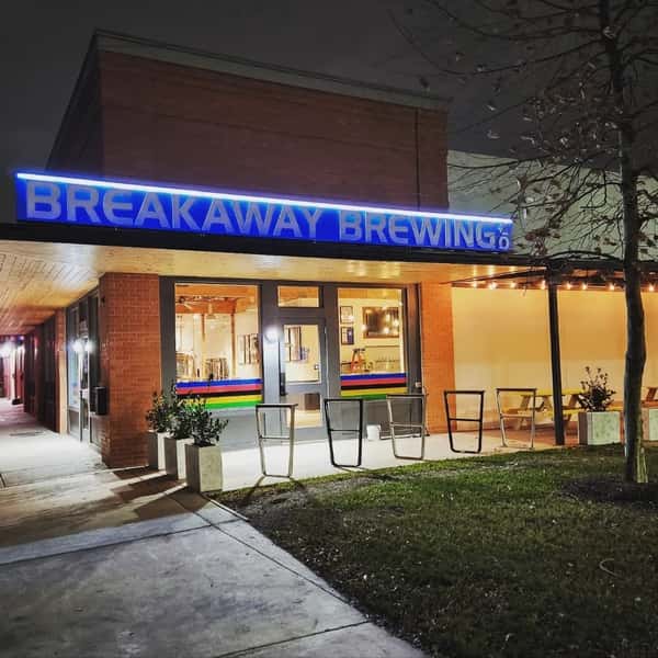 Breakaway Brewing storefront at night with bright blue neon sign, outdoor seating, and warm lights glowing inside.