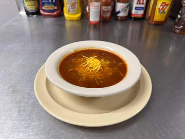 Bowl of chili topped with shredded cheddar cheese, served on a white plate with condiments in the background.
