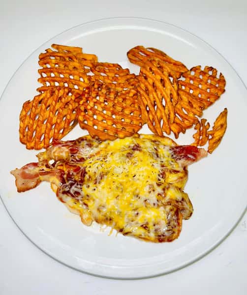 BBQ chicken breast topped with melted cheese and bacon, served with crispy waffle fries on a white plate.