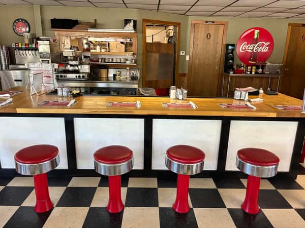 Classic diner counter with red swivel stools, a wooden countertop, Coca-Cola decor, and an open view into the kitchen area.