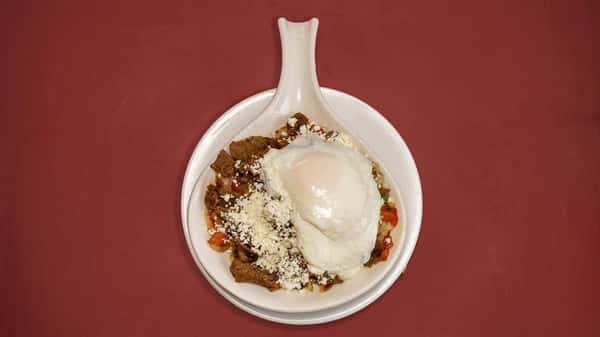 Chilaquiles topped with a poached egg, crumbled cheese, and sautéed vegetables, served in a white ceramic dish on a red tabletop.