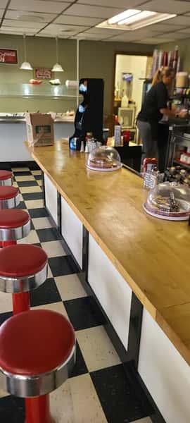 Retro diner counter with red stools, black-and-white checkered floor, and desserts under glass domes beside a working server.