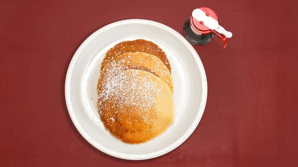 Stack of golden pancakes dusted with powdered sugar, served with a bottle of syrup on a red table.