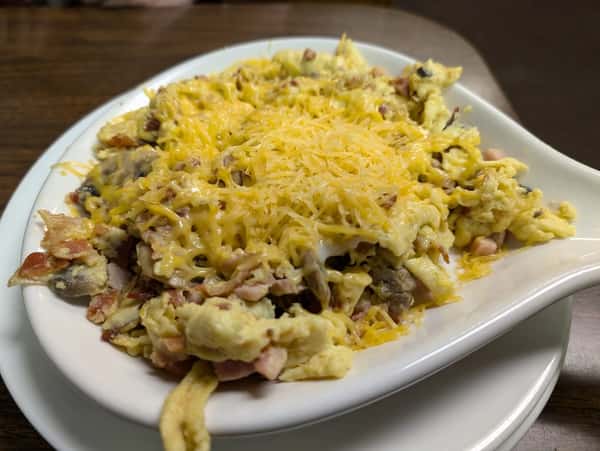 Scrambled eggs mixed with bacon, sausage, and vegetables, topped with shredded cheese, served in a white dish.