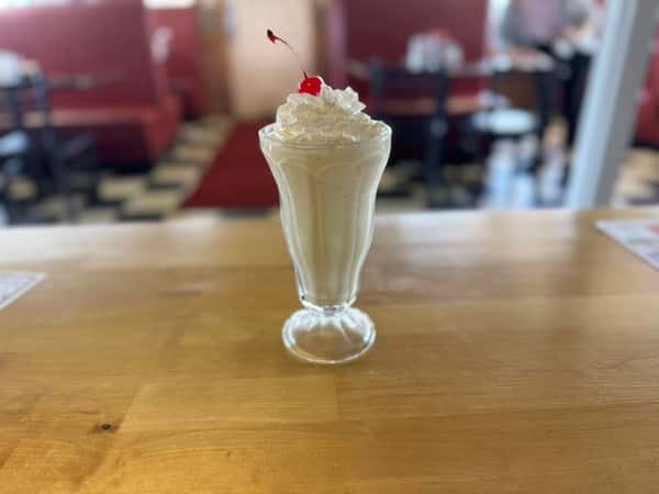 Vanilla milkshake topped with whipped cream and a cherry, served in a tall glass on a wooden counter in a diner setting.