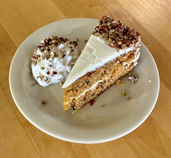 Slice of carrot cake with cream cheese frosting and chopped pecans, served with whipped cream topped with more pecans.