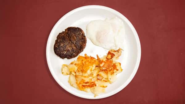 Classic breakfast plate with a hamburger patty, hash browns, and a sunny-side-up egg on a white plate over a red tabletop.