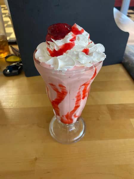 Strawberry milkshake swirled with syrup, topped with whipped cream and a cherry in a tall glass on a wooden table.
