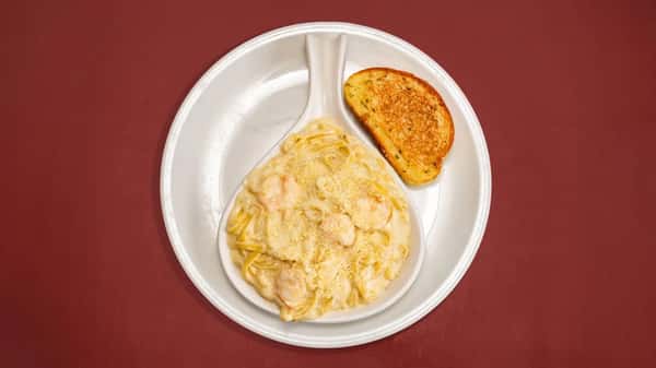 Shrimp fettuccine Alfredo topped with grated Parmesan, served with a slice of garlic toast on a white plate over a red tabletop.