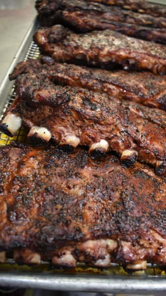 Several racks of slow-smoked BBQ ribs with a dark, peppery seasoned bark and charred bone ends, resting on a metal wire cooling rack.