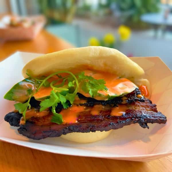 A close-up photograph of a fluffy, white steamed bao bun split open and served in a white paper food tray on a wooden table. The bun is generously filled with a thick slice of dark, smoked, charred pork belly. It is topped with bright orange sriracha aioli, fresh green cilantro sprigs, and green dill pickle slices. The background shows a gently blurred warm restaurant setting.