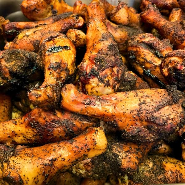 A close-up, mouth-watering shot of a massive pile of freshly smoked chicken drumsticks. The chicken has a rich, roasted golden-brown color with dark, charred edges, and is generously coated in a dark, savory, peppery dry rub.