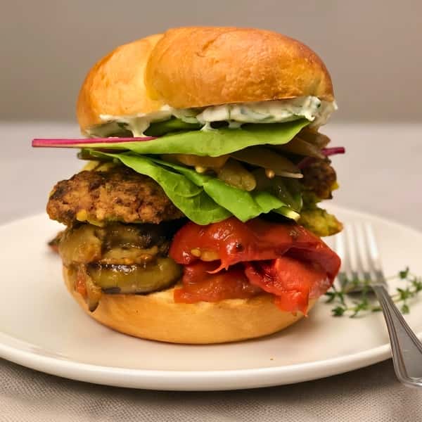 A close-up photograph of a hearty chickpea fritter sandwich served on a white round plate. A silver fork and a sprig of fresh thyme rest on the plate beside the sandwich. The sandwich is stacked high on a toasted brioche bun with roasted red peppers, grilled eggplant, a crispy chickpea patty, mixed greens, pickled red onion slices, and white cucumber yogurt sauce.
