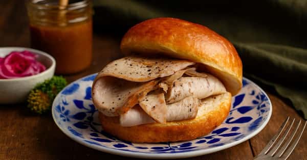 A close-up photograph of a smoked, hand-carved turkey breast sandwich generously filled with peppered turkey slices on a glossy brioche bun. The sandwich rests on a decorative blue and white patterned porcelain plate. In the background on a rustic wood table, there is a small bowl of vibrant pickled red onions and microgreens, a glass jar of rich BBQ sauce with a wooden honey dipper, and a silver fork. The setting is on a dark wood table with a dark green fabric in the background.