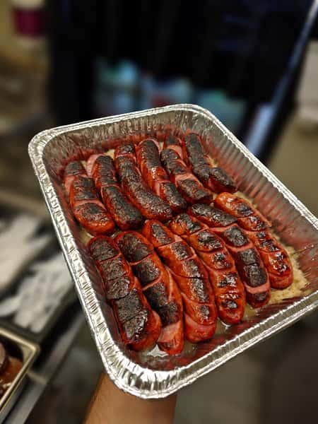 A close-up photograph showing a rectangular foil platter, held up by a person's hand, filled with multiple scored and grilled Bratwurst sausages resting on a bed of sauerkraut. The sausages are a rich reddish-brown, with charred, crisscrossed grill marks along their length.