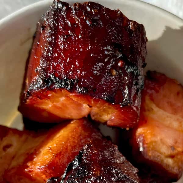 A macro shot of several smoked pork belly burnt ends piled in a bowl. The focus is on a large, glistening burnt end with a sticky, dark bark and a pinkish smoke ring visible on the edge of the tender meat.
