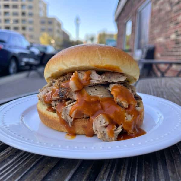 A close-up of a juicy pulled pork sandwich piled high on a toasted bun and generously smothered in a tangy orange-red BBQ sauce. The sandwich sits on a white beaded plate resting on a rustic wooden outdoor table. In the softly blurred background, a brick building and a sunny, tree-lined street scene in Arlington Heights can be seen.