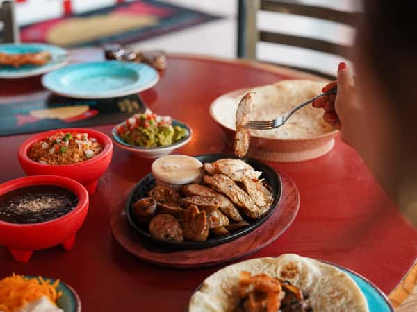 Mexican dining table featuring grilled meats, tortillas, salsa, guacamole, and side dishes.