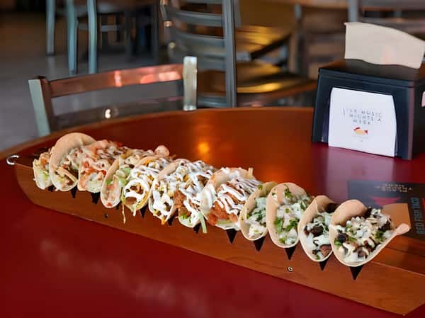 Long platter of tacos topped with fresh vegetables, cheese, and sauce.