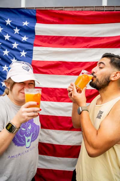 Two friends enjoying hazy craft beers in front of a large American flag at a festive brewery celebration in San Antonio.