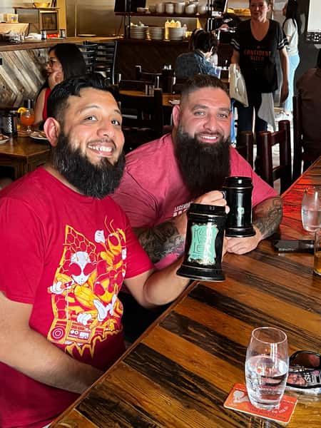 Two friends raising ceramic beer steins and smiling at a wooden table inside Künstler Brewing's rustic dining area.