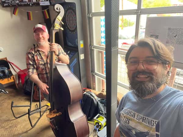 Two musicians smiling before a live set at Künstler Brewing, one holding an upright bass beside a dartboard and open garage door.