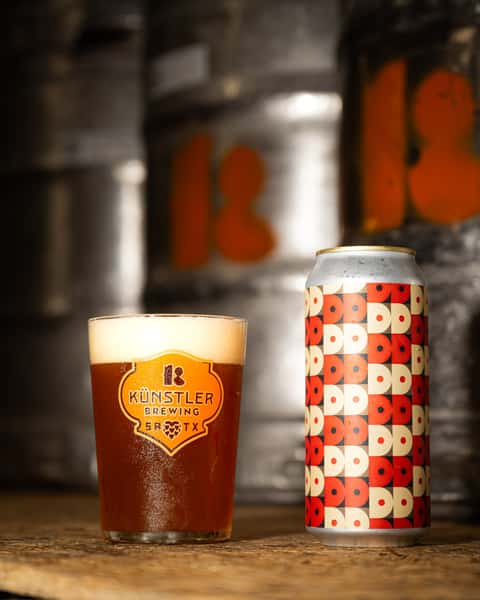 Branded pint glass filled with amber craft beer next to a stylish, patterned Künstler Brewing can, set in front of metallic beer kegs.