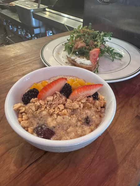 Healthy breakfast bowl of oatmeal topped with fresh berries, sliced strawberries, oranges, and granola, served alongside avocado toast with arugula and tomato.