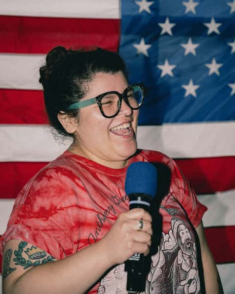 Smiling woman with a microphone standing in front of an American flag backdrop, enjoying open mic night or live entertainment at the brewery.