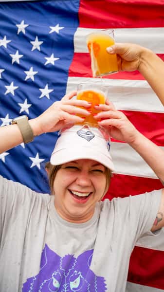 Woman laughing while balancing hazy craft beer glasses in front of a large American flag backdrop at a festive brewery event.