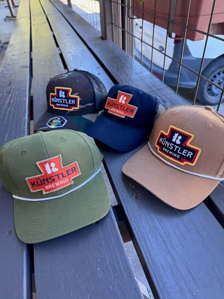 Künstler Brewing branded hats in green, navy, tan, and camo styles displayed on a wooden outdoor bench at the brewery's patio.