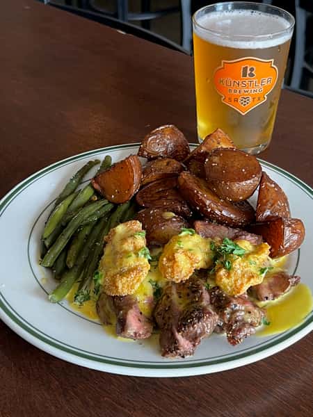 Plate of grilled steak with fried shrimp and hollandaise sauce, roasted potatoes, and green beans, paired with a pint of Künstler Brewing craft beer.