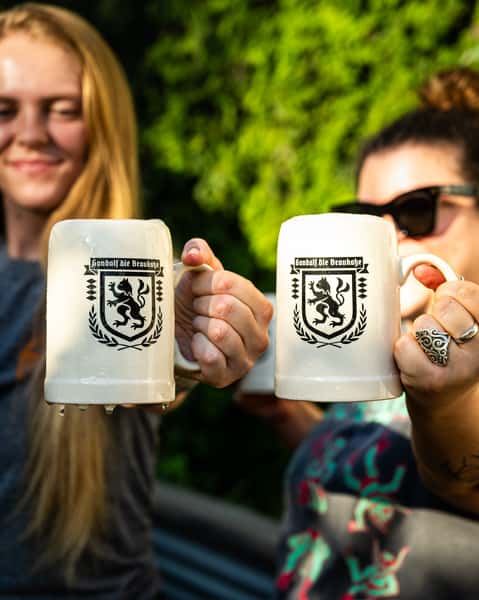 Two women holding ceramic German-style beer steins featuring a heraldic lion crest, enjoying a sunny afternoon at Künstler Brewing.
