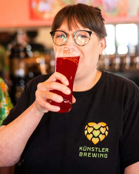 Woman enjoying a vibrant red fruit beer in a branded glass at Künstler Brewing, wearing a logo tee with a hop heart design, inside the cozy and colorful brewery taproom.