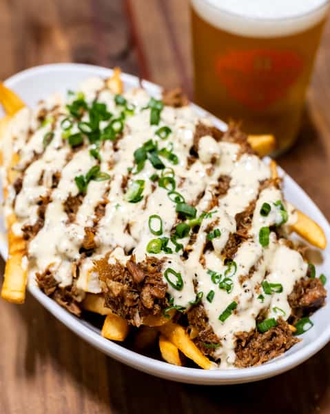 Close-up of crispy French fries topped with shredded beef, creamy white sauce, and green onions, served with a pint of Künstler Brewing craft beer.