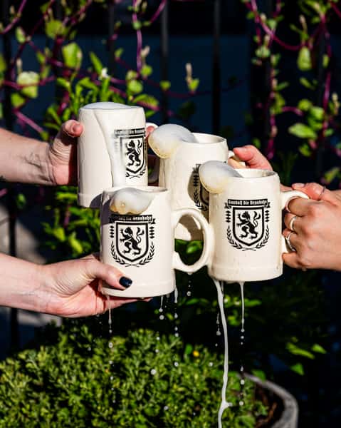 Close-up of four hands toasting ceramic beer steins overflowing with frothy foam at a German-style beer garden, with greenery and climbing vines in the background. The steins feature a black lion crest with the text "Gambol die Braubok," evoking a festive Bavarian or Oktoberfest ambiance.