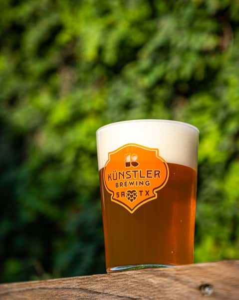 Künstler Brewing pint glass filled with golden amber craft beer, topped with frothy foam, set on a wooden ledge with leafy greenery in the background.