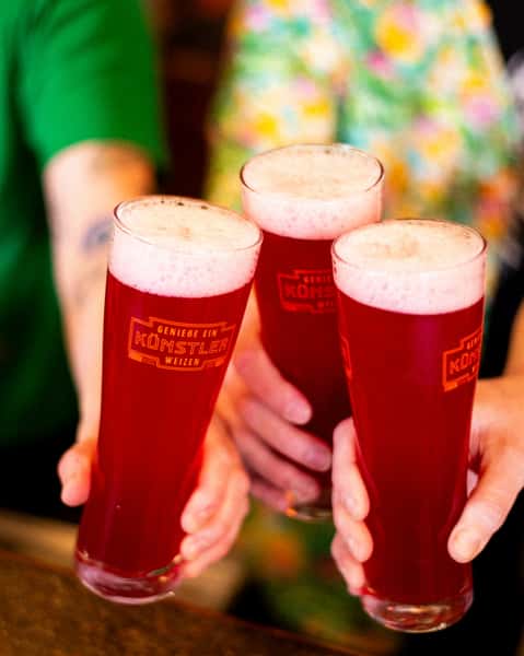 Close-up of three tall weizen glasses filled with ruby-hued fruit beer from Künstler Brewing, topped with a frothy head.